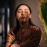 A woman embraces herself in serene indoor lighting with shadow play on her face.